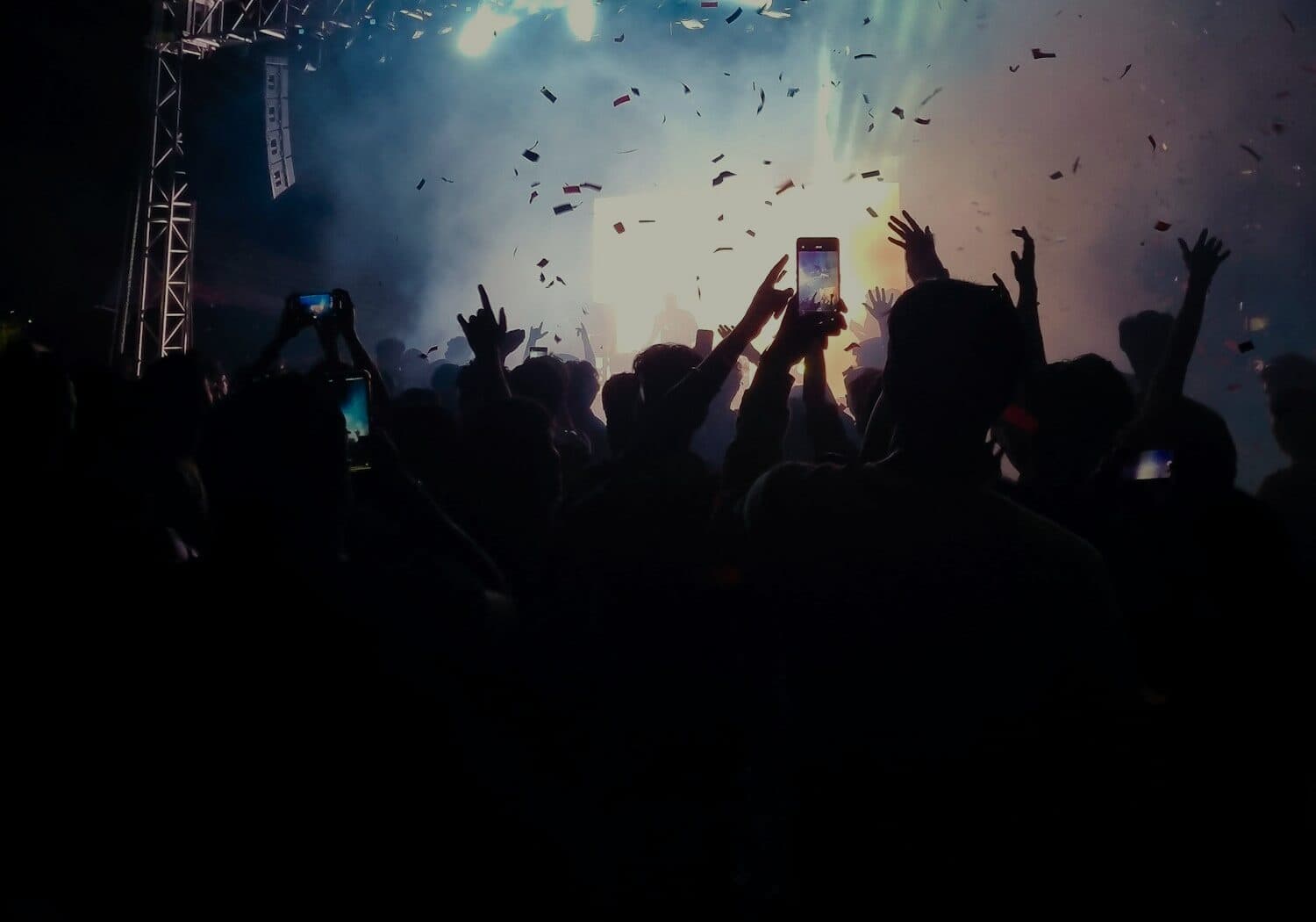 An audience in silhouette from behind, hands and phone screens raised toward a lit stage
